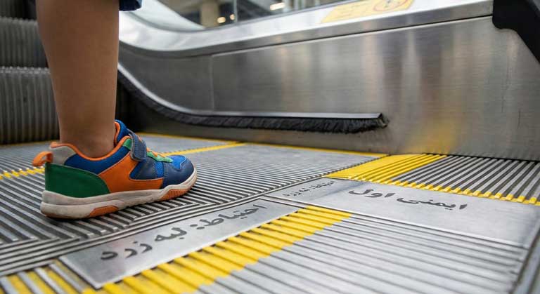 پای کودک روی پله برقی در محدوده ایمن خطوط زرد و دور از دیواره کناری.