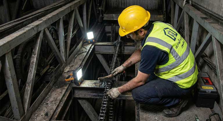 تکنسین پله برقی در حال تمیزکاری زنجیر و آمادهسازی برای روغن کاری در موتورخانه پایین.